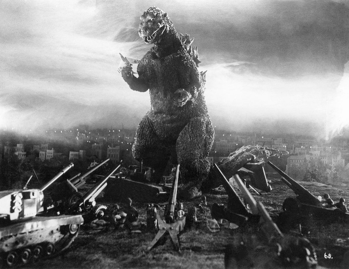 An image of Godzilla from the original 1954 film, with cannons being pointed at him as he destroys Tokyo.